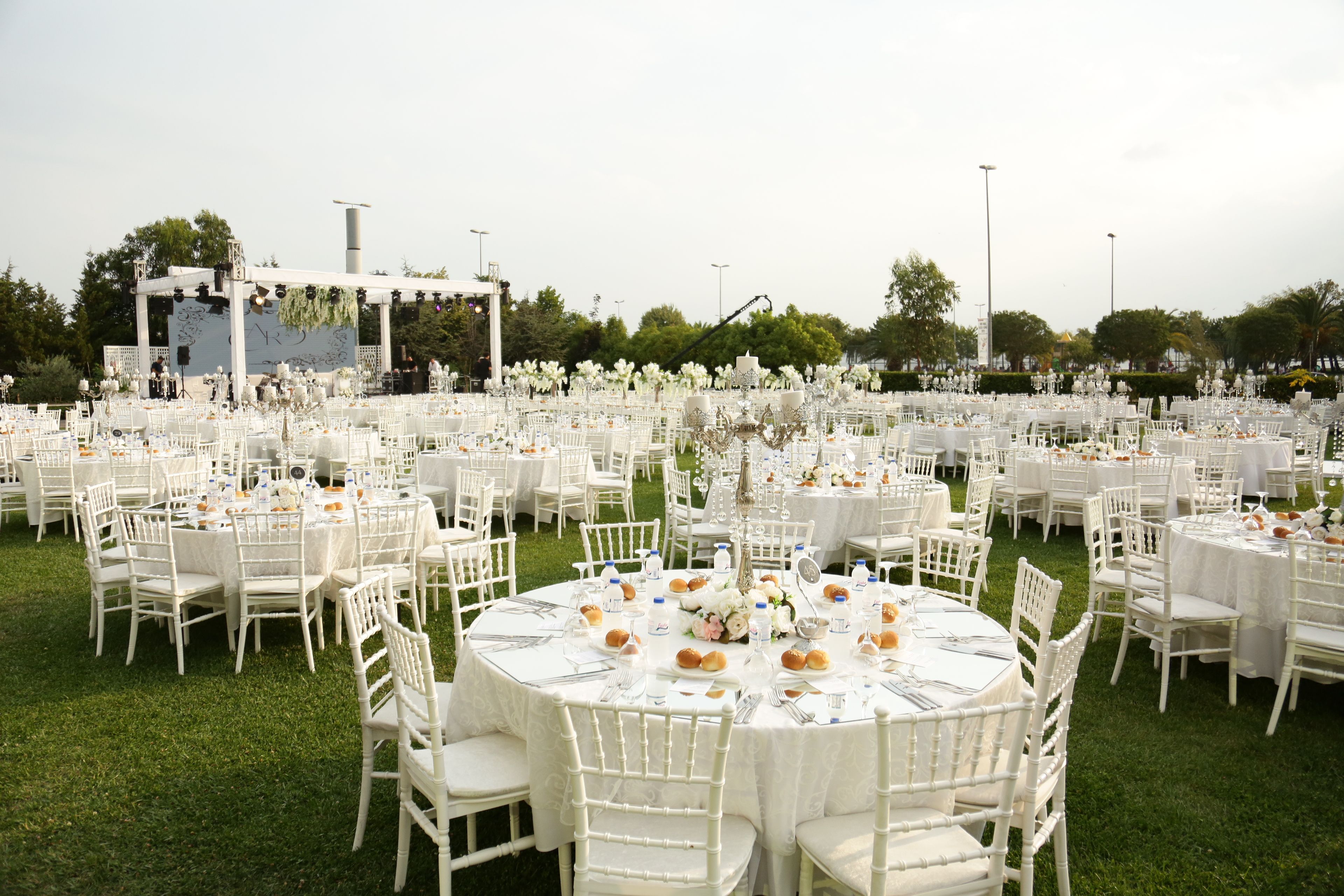 An outdoor event setup with numerous white round tables and chairs on a green lawn.